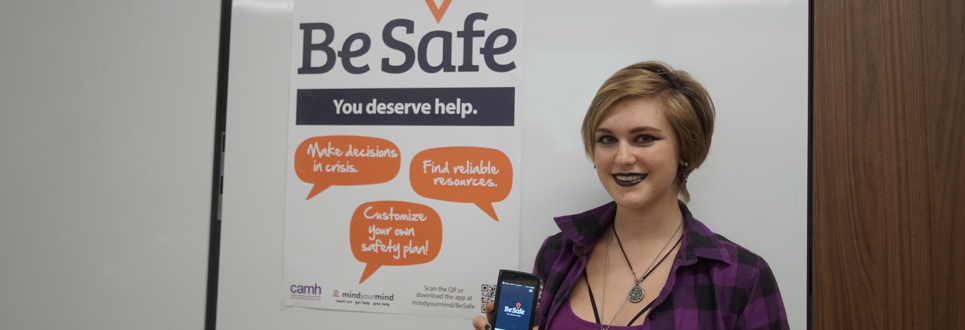 Female student holding cell phone with Be Safe App and Be Safe poster in background